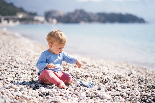 Little kid in a blue blouse and red pants sits on a pebble beach holding a stick in his hand