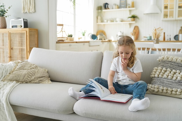 Little kid girl preschooler read fairytale book sitting on cozy couch at home alone Child education