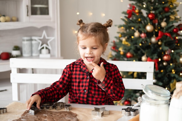 little kid girl in red pajama cooking festive gingerbread in christmas decorated kitchen