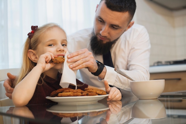 Little kid and her father eating biscuits at the kitchen while father is cleaning her face. Father and daughter in the morning at kitchen eating.