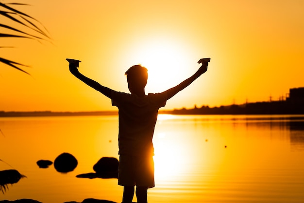 Little kid holds two paper planes in spread hands and plays at sunset on the lake background Time outdoor Black silhuette against sundown