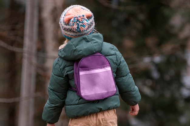 Little kid in knitted hat and jacket wears small backpack in pine forest Child in fall in woods back view