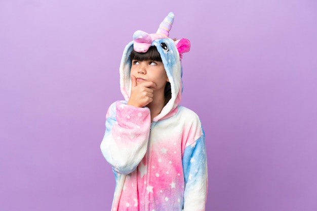 Little kid wearing a unicorn pajama isolated on purple background having doubts and with confuse face expression