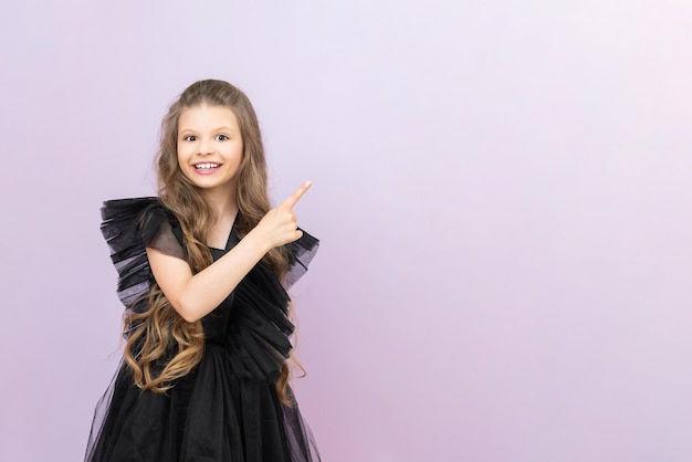 The little lady points to your advertisement A beautiful little girl with curly hair in a black dress on a pink isolated background Copy space