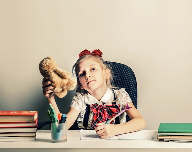 A little lefthanded girl in a school uniform is sitting at a table with books and pens thinking what to write in her notebook