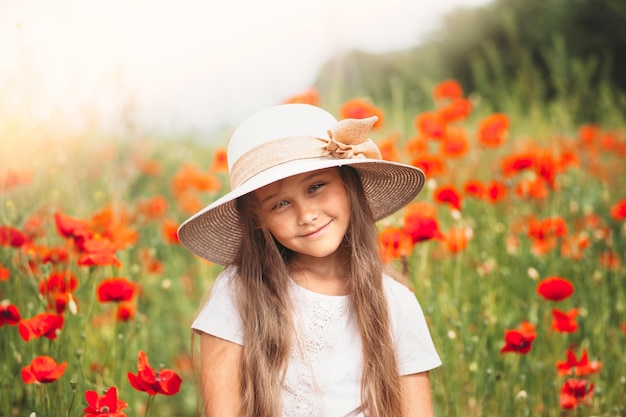 Little longhaired girl in hat posing at field of poppies with on summer sun