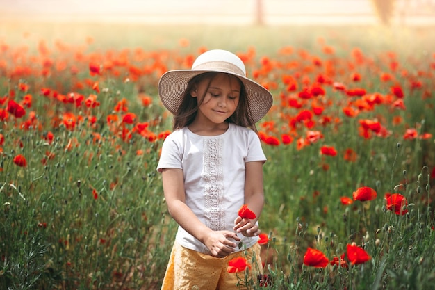 Little longhaired girl in hat posing at field of poppies with on summer sun