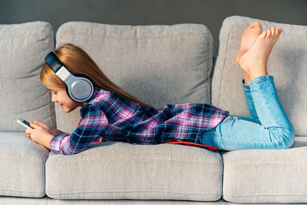 Photo little music lover. beautiful little girl in headphones holding her smart phone with smile while lying on front on the couch at home