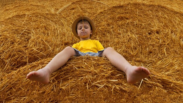 Little nice boy dressed in a yellow shirt and denim shorts and modern hat sits on a haystack