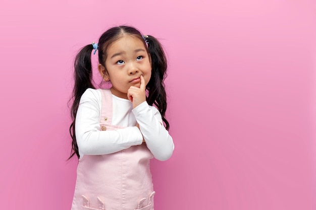 little pensive asian girl in pink sundress looks away and dreams on pink isolated background