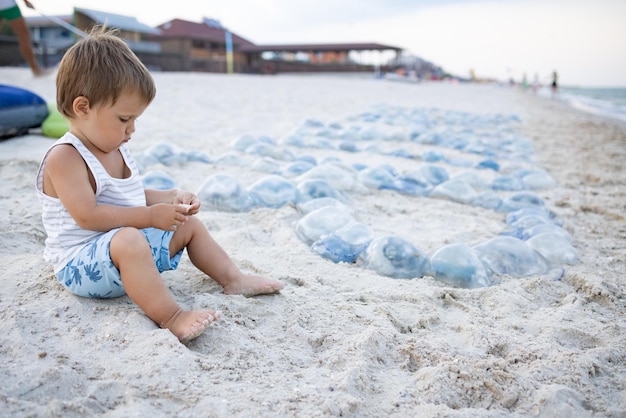 Little pensive wary boy is playing with seashells and warm yellow sand while sitting on a large spacious beach, strewn with dangerous poisonous blue jellyfish near the calm summer sea