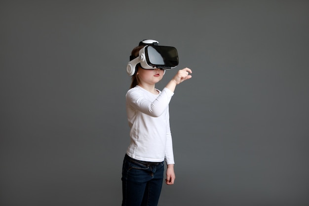 Little preschool girl experiencing virtual reality eyeglassses in white long sleeve shirt over grey surface.