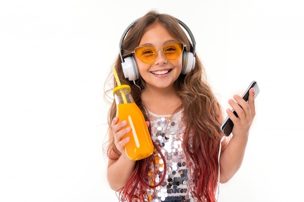 Little pretty caucasian girl listen to music with big earphones and drinks orange juice, picture isolated on white wall