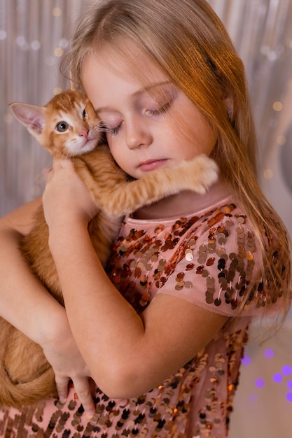 A little princess girl in an elegant dress holds a red kitten in her hands. The symbol of the year