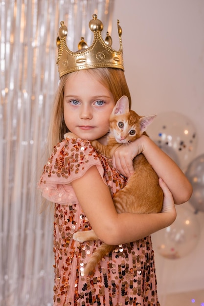 A little princess girl in an elegant dress holds a red kitten in her hands. The symbol of the year