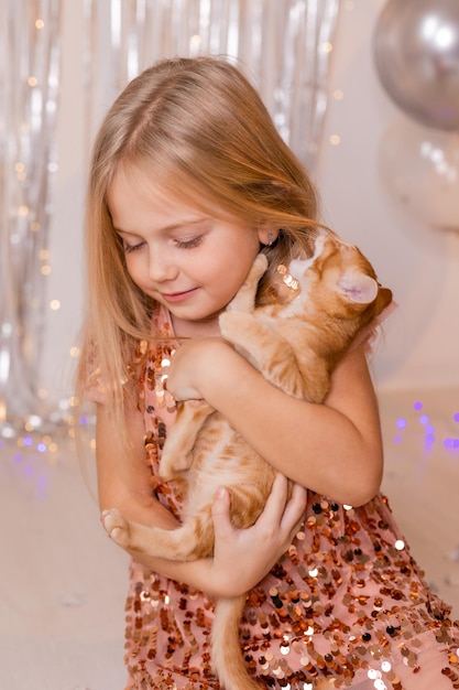 A little princess girl in an elegant dress holds a red kitten in her hands. The symbol of the year