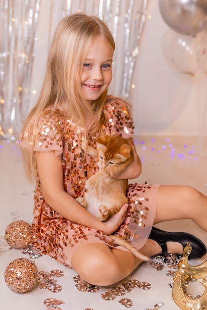 A little princess girl in an elegant dress holds a red kitten in her hands. The symbol of the year