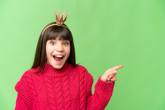 Little princess with crown over isolated chroma key background surprised and pointing finger to the side