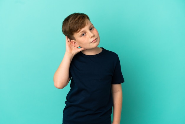 Little redhead boy isolated on blue surface listening to something by putting hand on the ear