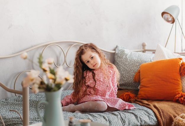 A little romantic girl with long hair sits on her bed in the children's bedroom