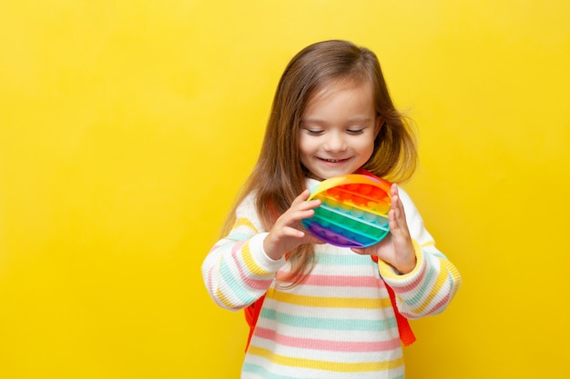 A little schoolgirl in a striped blouse and with a backpack holds a pop it in her hands and smiles