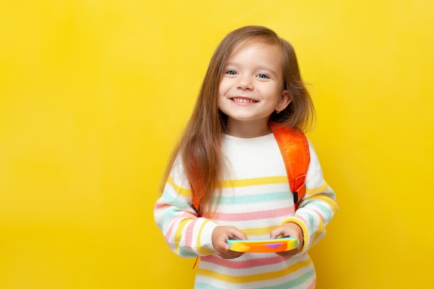 A little schoolgirl in a striped blouse and with a backpack holds a pop it in her hands and smiles