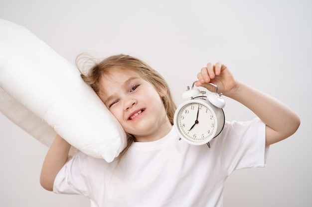 A little sleepy girl hugs a pillow and an alarm clock on a white background. 