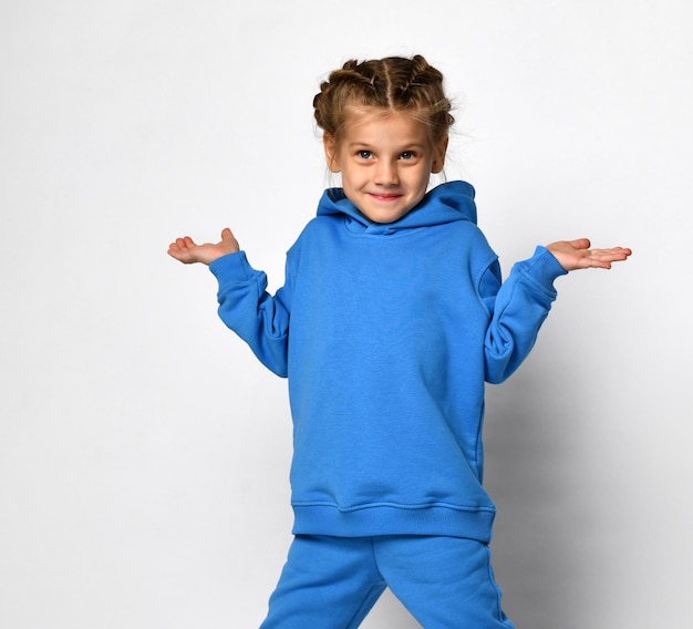 Little sly girl in a blue sports suit shrugs while standing on a white background