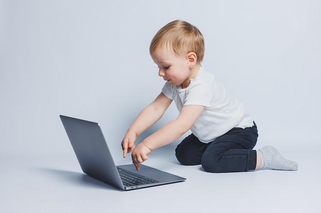 Little smart boy 34 years old sits with a laptop on a white background A child in a white Tshirt and black trousers sits at a laptop and looks at the screen Modern progressive children