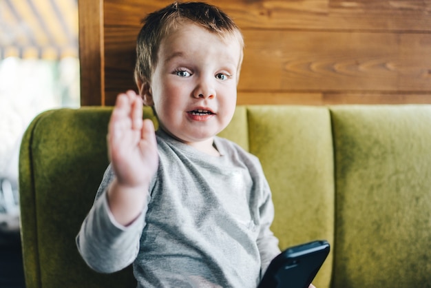 Little smiling baby boy sitting on sofa with mobile phone in hand and waving