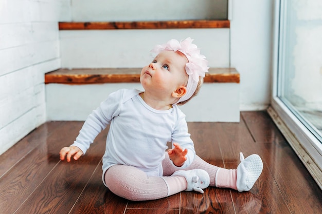 Little smiling baby girl one year old wearing spring wreath siting on floor in bright light living room near window and playing with gerbera flowers Happy kid playing at home Childhood concept