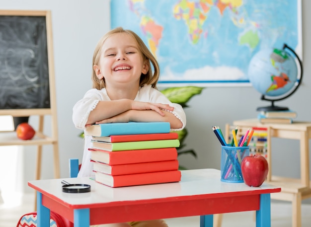 Little smiling blond girl sitting at the white desk and holding hands on the books in the spacious school class