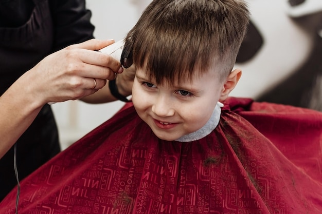 Little smiling boy cut with hairdresser's machine Closeup of woman hands grooming kid boy hair in barber shop Portrait of male child at the barber shop to cut his hair