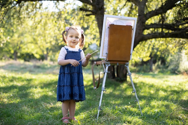 Little smiling girl in jeans dress, painting at green summer park outdoor. Talented small girl artist, holding brush in hands, posing  near the easel at sunny spring garden.