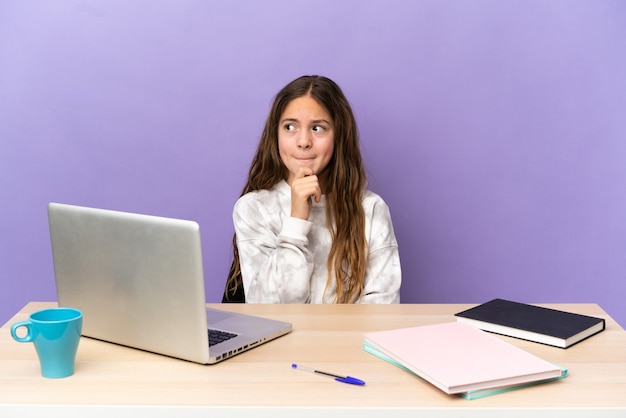 Little student girl in a workplace with a laptop isolated on purple background having doubts and thinking