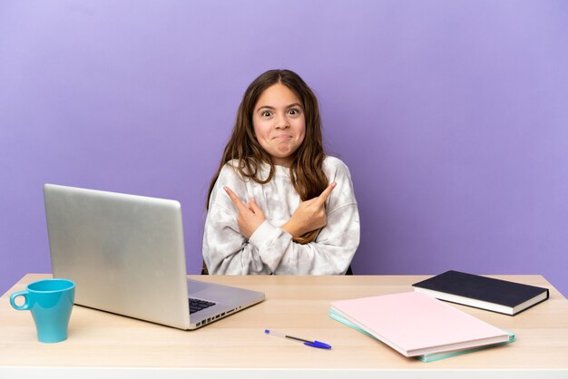 Little student girl in a workplace with a laptop isolated on purple background pointing to the laterals having doubts