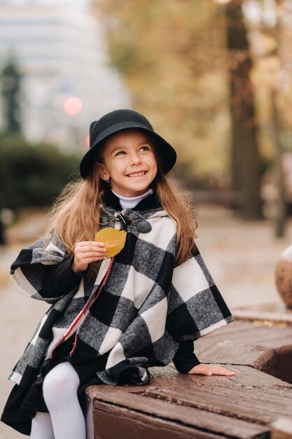 A little stylish girl in autumn clothes is sitting on a bench in the autumn city