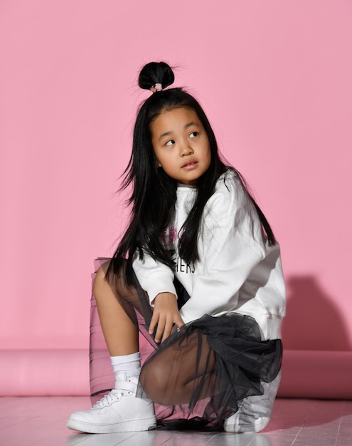 Little stylish girl model posing sitting on a pink background in the studio looking up Girl wearing a black skirt white sweater and sneakers Concept of stylish clothes for schoolchildren