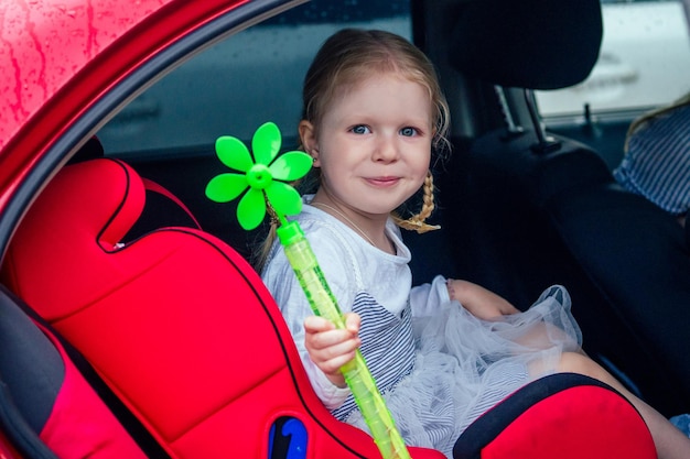 Little and sweet blonde girl in a cute gray dress beautiful sitting in the red car child seat seat for childrenin.