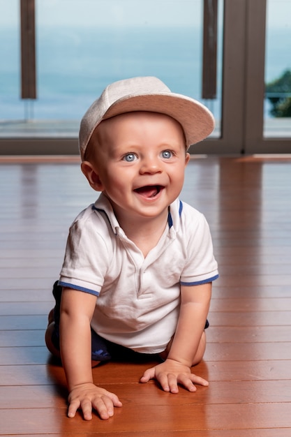 Little toddler boy in gray fashion cap and barefoot, crawling on the floor, smiling and looking to the side