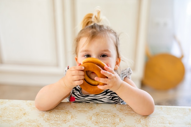 Little toddler kid having lunch in the warm sunny kitchen. Blonde girl with funny ponytail playing with two tasty bagels