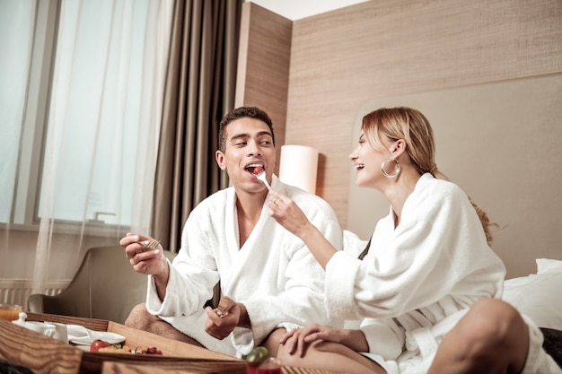 Little tomato. Dark-eyed husband biting piece of little tomato having breakfast with his appealing stylish wife