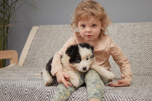 Little wavy haired girl happily playing with adorable puppy on cozy sofa in her room enjoying spendi