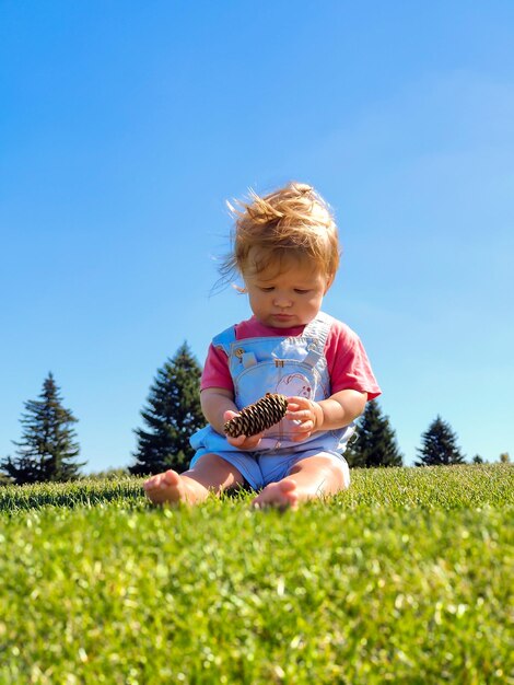 A little white boy is sitting on the green grass against the background of the blue sky and playing