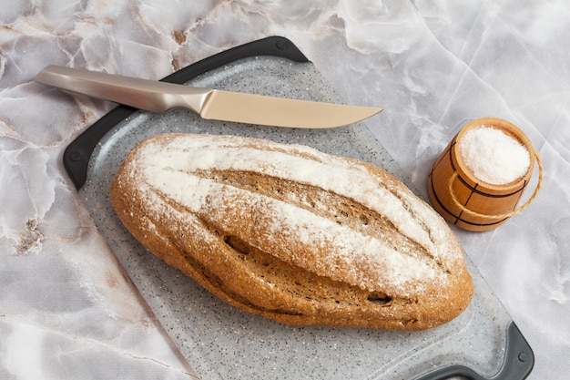 Loaf of rye bread and a kitchen knife on a cutting board, a wooden salt cellar with the gray background. Top view.