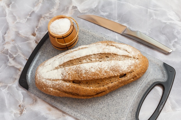 Loaf of rye bread and a wooden salt cellar on a cutting board, a kitchen knife with the gray surface