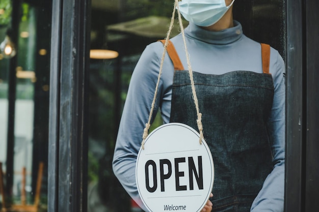 Lockdown. waitress staff woman wearing protection face mask turning OPEN sign board on glass door in modern cafe coffee shop, cafe restaurant, retail store, small business, food and drink concept