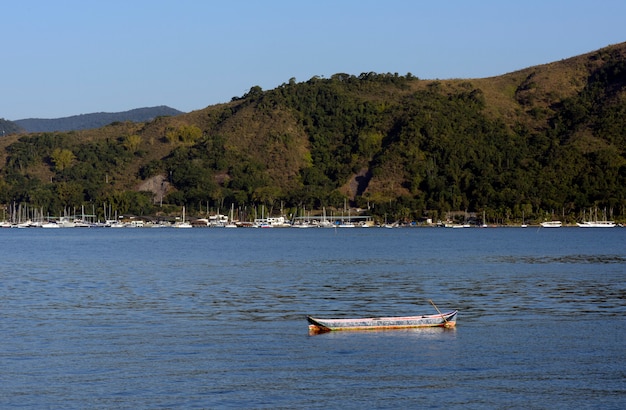 Lone canoe on calm sea, with boats, hill and blue sky