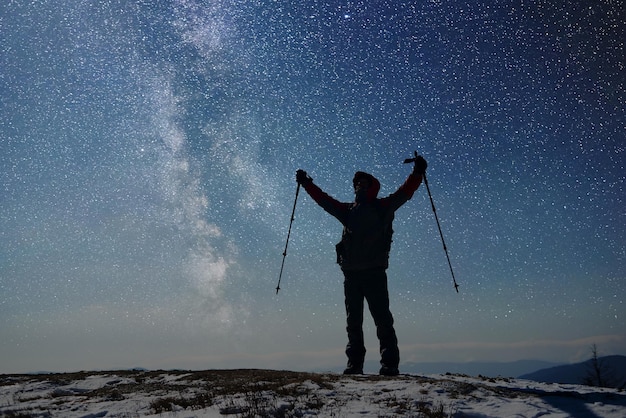 Lone silhouette of adult male hiker on top of snowcapped mountain in anticipation of dawn Starry sky on background