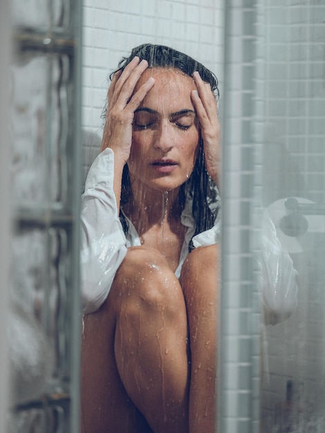 Lonely brunette with closed eyes in white wet shirt sitting in tiled shower under water and touching head gently